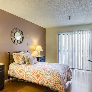A cozy bedroom with a well-decorated bed and warm lighting, featuring a decorative wall clock and large window blinds allowing natural light.
