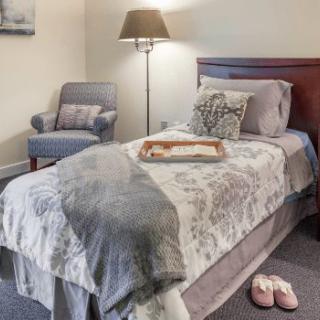 A cozy bedroom with a single bed adorned with patterned bedding, a decorative pillow, and a tray. A pair of pink slippers rests beside the bed, accompanied by a comfortable chair and lamp.