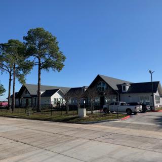 A modern building with a sloped roof, surrounded by tall trees and a parking lot under a clear blue sky.