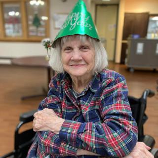 A cheerful elderly woman wearing a green party hat, smiling while seated in her wheelchair.
