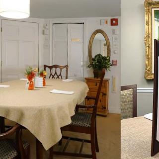 A cozy dining room with a round table covered by a beige tablecloth, surrounded by wooden chairs, featuring decorative elements like a mirror and a framed painting.