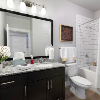 A modern bathroom with dark cabinetry and a marble countertop, featuring a large mirror and a bathtub with white subway tiles.