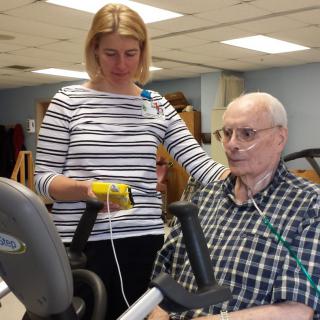 An elderly man with an oxygen tube exercises on a stationary bike while being assisted by a healthcare worker.