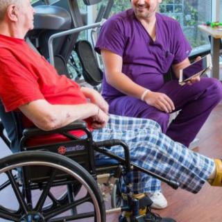 A nurse in purple scrubs converses with an elderly patient seated in a wheelchair, fostering a supportive and caring environment.