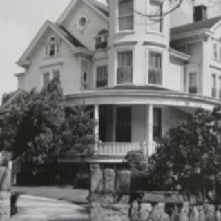 A black and white photograph of a large Victorian-style house with a prominent porch.