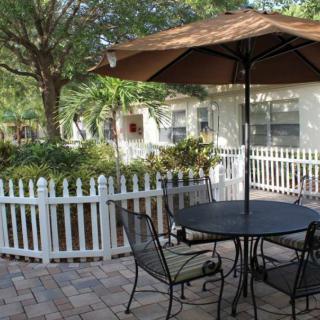 A cozy outdoor patio area with a table, chairs, and an umbrella, surrounded by a white picket fence and lush greenery.
