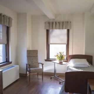 A hospital room with a bed, chair, and flowers on a table by the window.