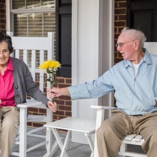 An elderly couple sitting on a porch, with one person giving flowers to the other.