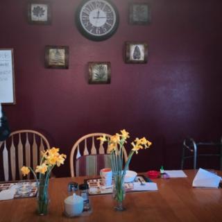 A dining room with a wooden table, chairs, and daffodils in vases, decorated with wall art and a clock.