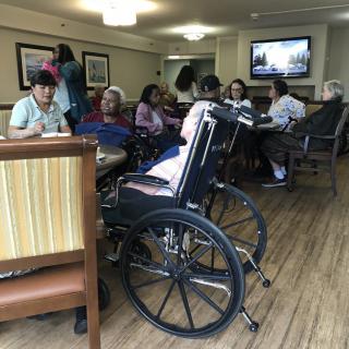 A group of people in a nursing home, some in wheelchairs, socializing in a common room.
