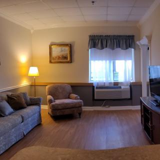 A cozy living room with a sofa, armchair, lamp, and television. The window is adorned with curtains and there is artwork on the wall.