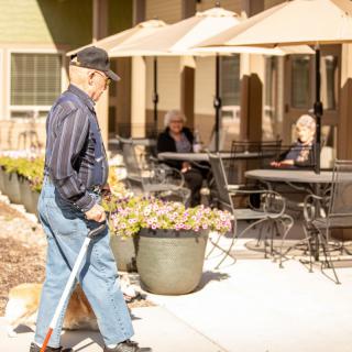 An elderly man walks with a cane past an outdoor patio area under sunny skies.