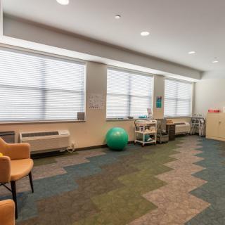 A spacious therapy room featuring chairs, a green exercise ball, and various equipment with large windows allowing natural light.