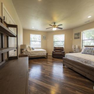 A spacious bedroom with wood flooring, featuring two beds, a ceiling fan, and multiple windows.