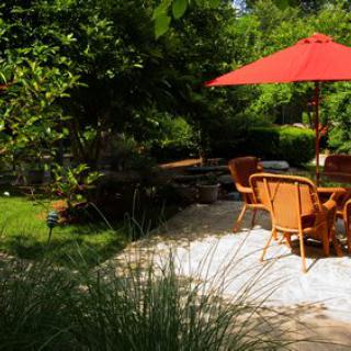 A serene garden patio with chairs and a red umbrella, surrounded by lush greenery.