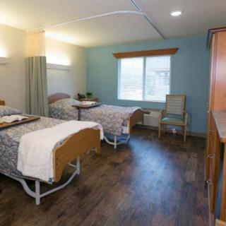 A hospital room with two patient beds separated by a curtain, featuring wooden flooring and a window providing natural light.