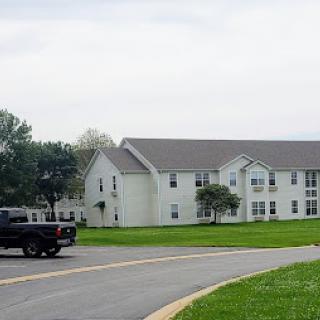 A two-story residential building surrounded by grass and trees, with a truck driving along the curved road.