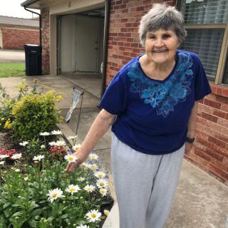 A smiling woman tends to her garden in front of a brick house.