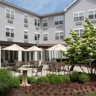 A well-maintained outdoor patio area with chairs, umbrellas, and lush landscaping in front of a multi-story building.