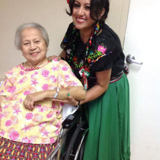 An elderly woman in a wheelchair is smiling while a person dressed in traditional clothing stands next to her.