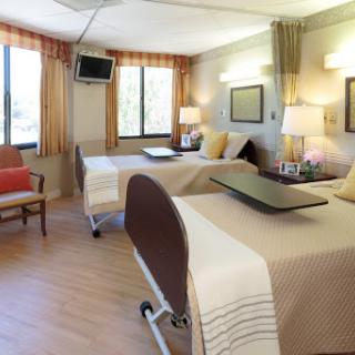 A spacious and well-lit hospital patient room with two beds, natural light from windows, and comfortable furnishings.