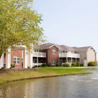 A scenic view of an apartment building by a tranquil lake, surrounded by greenery.