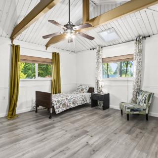 A spacious bedroom with a minimalist design, featuring wooden ceiling beams, a ceiling fan, and large windows letting in natural light.