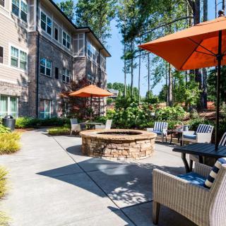 A cozy outdoor patio area with a fire pit, surrounded by chairs and orange umbrellas, set against the backdrop of an apartment building and lush greenery.