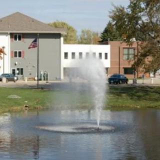 A scenic view of a fountain in a pond with buildings and trees in the background.