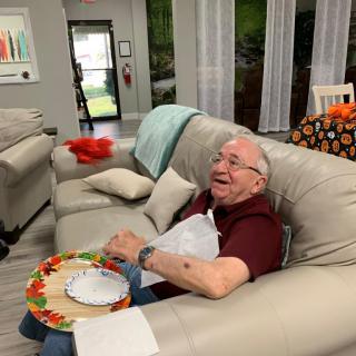 An elderly man sitting on a sofa in a living room, holding a plate and laughing.