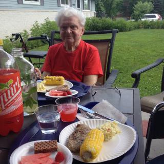 A person enjoying an outdoor meal with corn, watermelon, and soda on a sunny day.