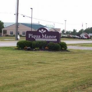 Piqua Manor sign in a grassy area with a building in the background.