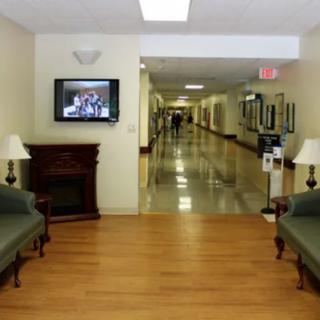 A hospital hallway with a waiting area, featuring sofas, a television, and a corridor leading further inside.