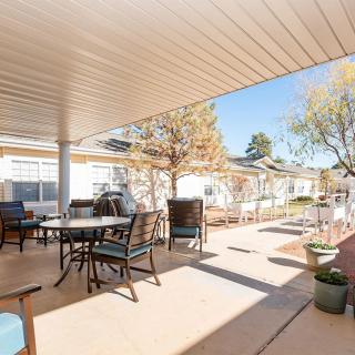 A sunny outdoor patio with tables and chairs under a covered area, surrounded by trees and plants.