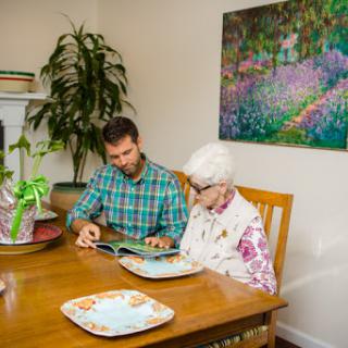 A young man shares a moment with an elderly woman at the dining table, engaging in a reading activity amidst a cozy home setting.