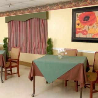 A cozy dining area with tables covered in green and brown tablecloths, surrounded by chairs. A decorative floral painting hangs on the wall, complemented by striped curtains and small potted plants.