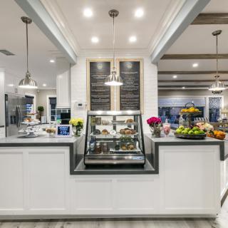 A modern cafe interior with a well-lit bakery display counter featuring various pastries, surrounded by vibrant flowers and a fruit basket.