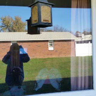 A reflection of a person standing by a window, with a view of an outdoor setting and a lantern hanging outside.