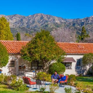 A charming home with a red-tiled roof set against a backdrop of mountains, featuring a lush garden and patio where people are relaxing.
