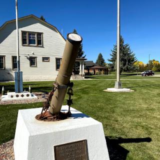 A small cannon monument on a pedestal in front of a building with flagpoles and green lawns under a clear blue sky.