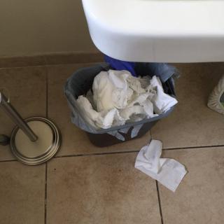 A bathroom scene with a trash can overflowing with tissue paper next to a sink.