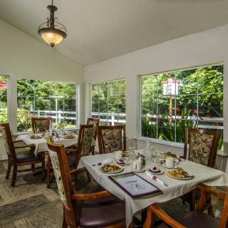 A cozy dining room with elegant tables and chairs, surrounded by large windows showcasing lush greenery.