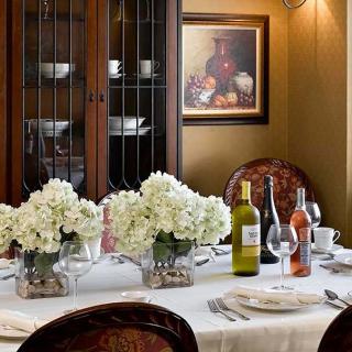 An elegantly set dining table with flowers and wine bottles, surrounded by ornate chairs, a decorative cabinet, and a framed painting on the wall.
