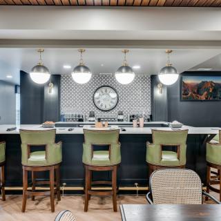 A modern bar interior featuring green stools, pendant lighting, and a decorative clock on a tiled wall.