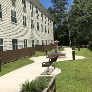 A pathway lined with benches next to a multi-story building, surrounded by trees and grass.