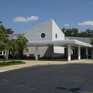 A modern building with a unique roof design and a parking lot in front, surrounded by trees on a clear day.