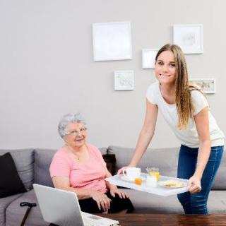 A caregiver serving a meal to a senior woman in a living room setting.