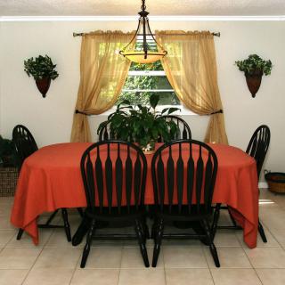 A dining room with a table covered in an orange tablecloth, surrounded by chairs and decorated with curtains and plants.
