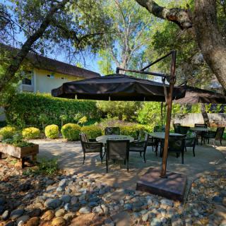 A tranquil outdoor seating area with tables and chairs shaded by an umbrella, surrounded by lush greenery and trees.