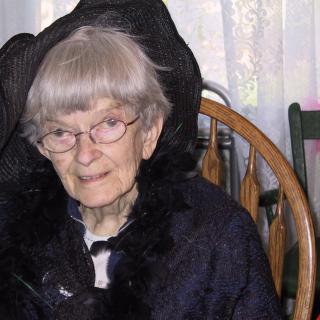 An elderly woman wearing glasses and a hat, sitting on a wooden chair indoors.